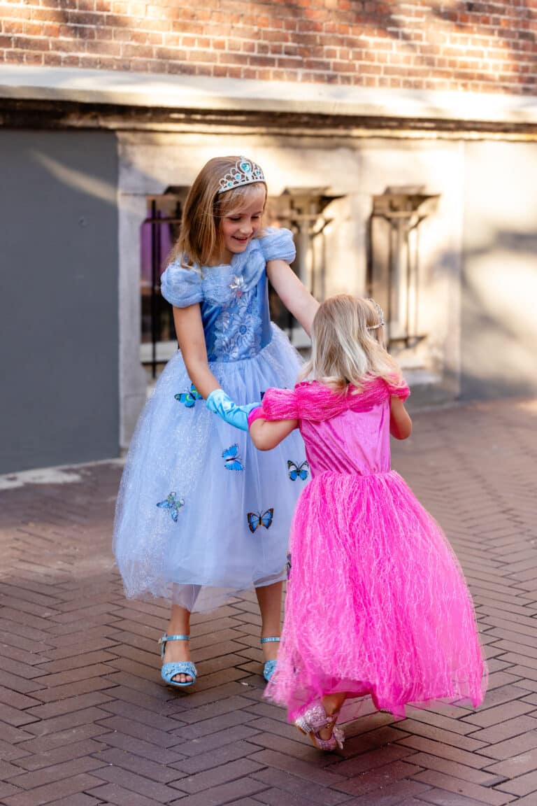 Meisje draagt een blauwe doornroosje jurk met vlinders en tiara, naast een kind in een roze prinsessenjurk.